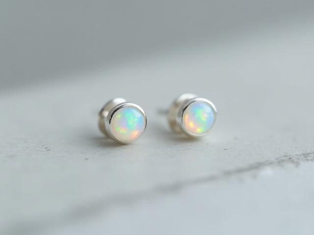 Pair of minimalist sterling silver stud earrings, each featuring a small, iridescent Australian opal, photographed on a smooth grey stone.